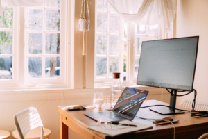 A white laptop and monitor on a table with clean windows allowing natural light to brighten the workspace.